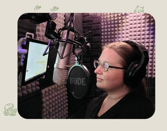 Picture of Tiffanie in her old sound booth, reading a script. there is a microphone and a wall mounted ipad in front of her. She is wearing a black shirt, glasses and headphones