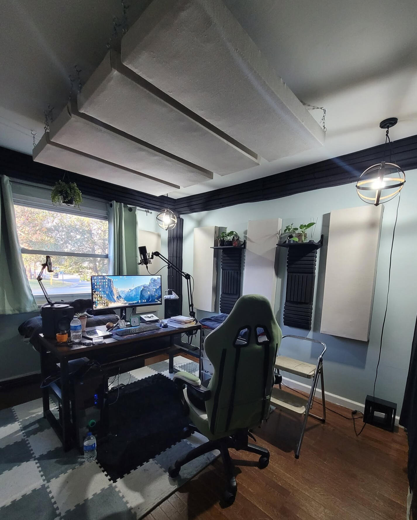 A sound treated studio with sage colored sound dampening curtains, Canvas colored sound panels, black base traps and thick acoustic foam, an desk 1/3 center placement with audio equipment for optimal sound.